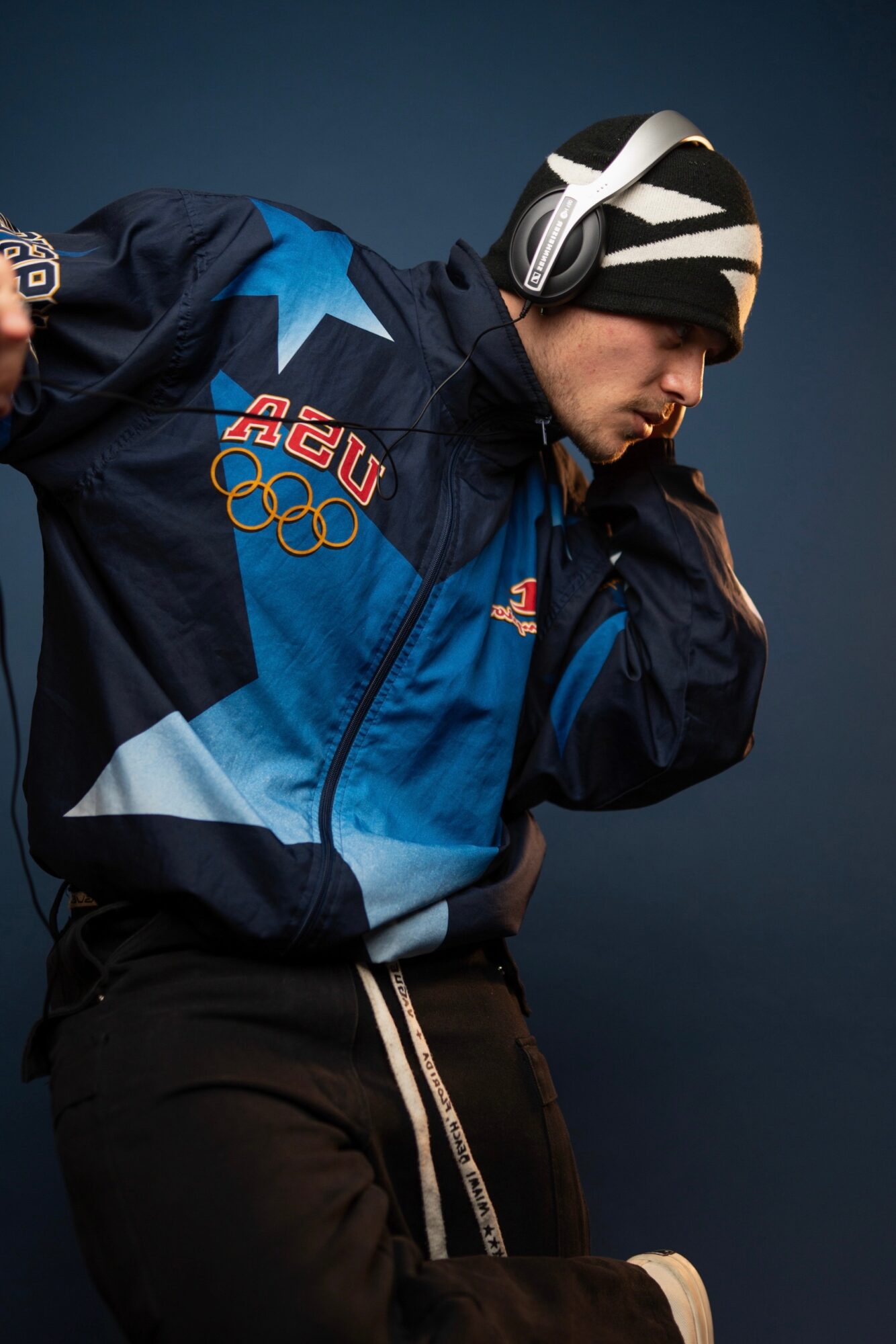 Athlete wearing a blue jacket with Olympic logos, headphones, and a helmet, preparing for a winter sport, against a dark background.