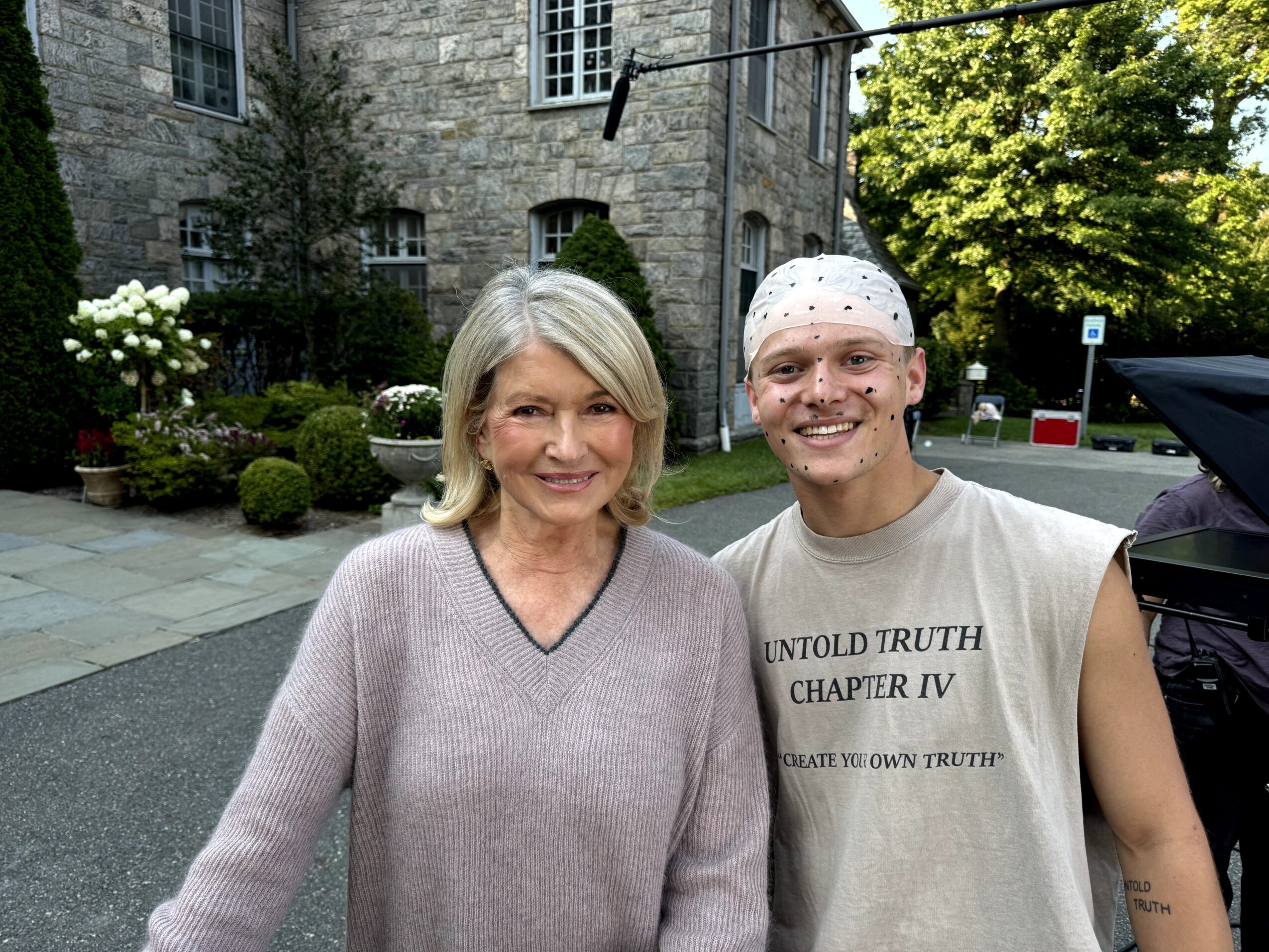 A woman and a young man stand outdoors in front of a stone building with trees and bushes, smiling at the camera.