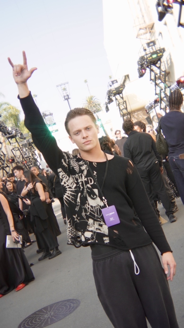 Young man with short hair raising his right hand in a peace sign at an outdoor event, crowd and stage in background.