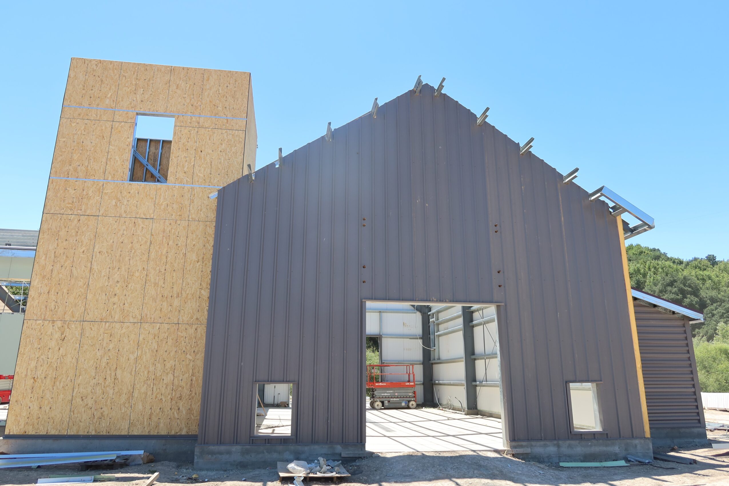 Partially constructed building with a black metal exterior and a wooden section, open garage door, and small windows, under a blue sky.