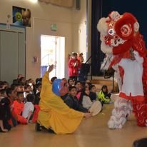 Children watching a lion dance performance indoors, with a performer in a red and white lion costume. Audience seated on the floor.