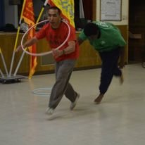 Two people playing with hula hoops indoors, one in red shirt and gray pants, the other in green shirt and dark pants, on a wooden floor.