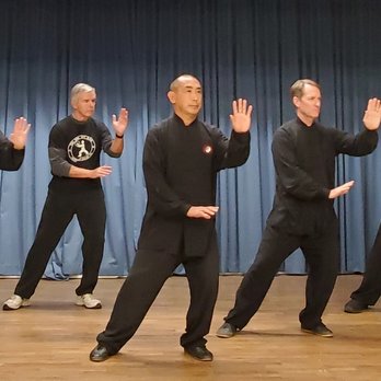 Four people practicing martial arts or Tai Chi on stage with blue curtains, wearing black clothing, performing synchronized movements.