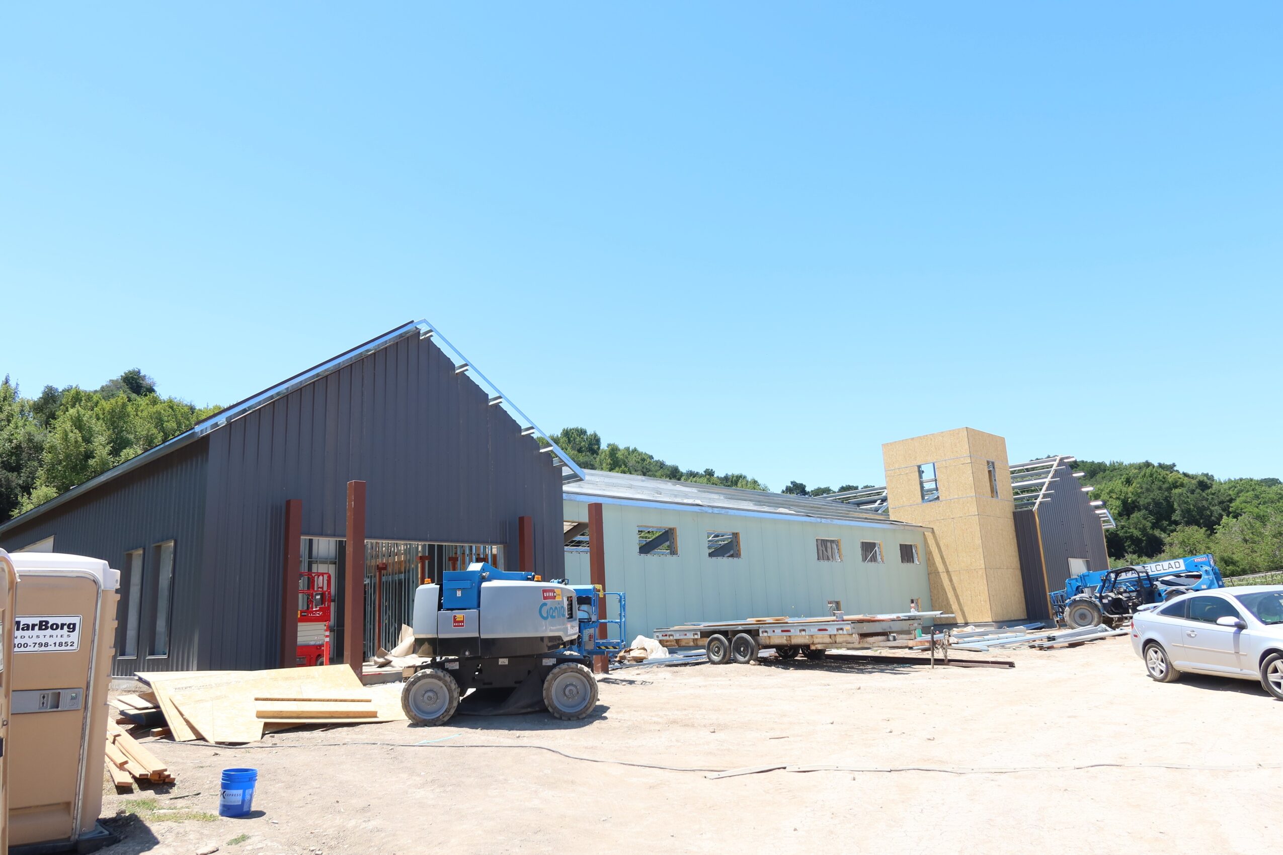 Construction site with a gray building, construction equipment, and a car under a clear blue sky.