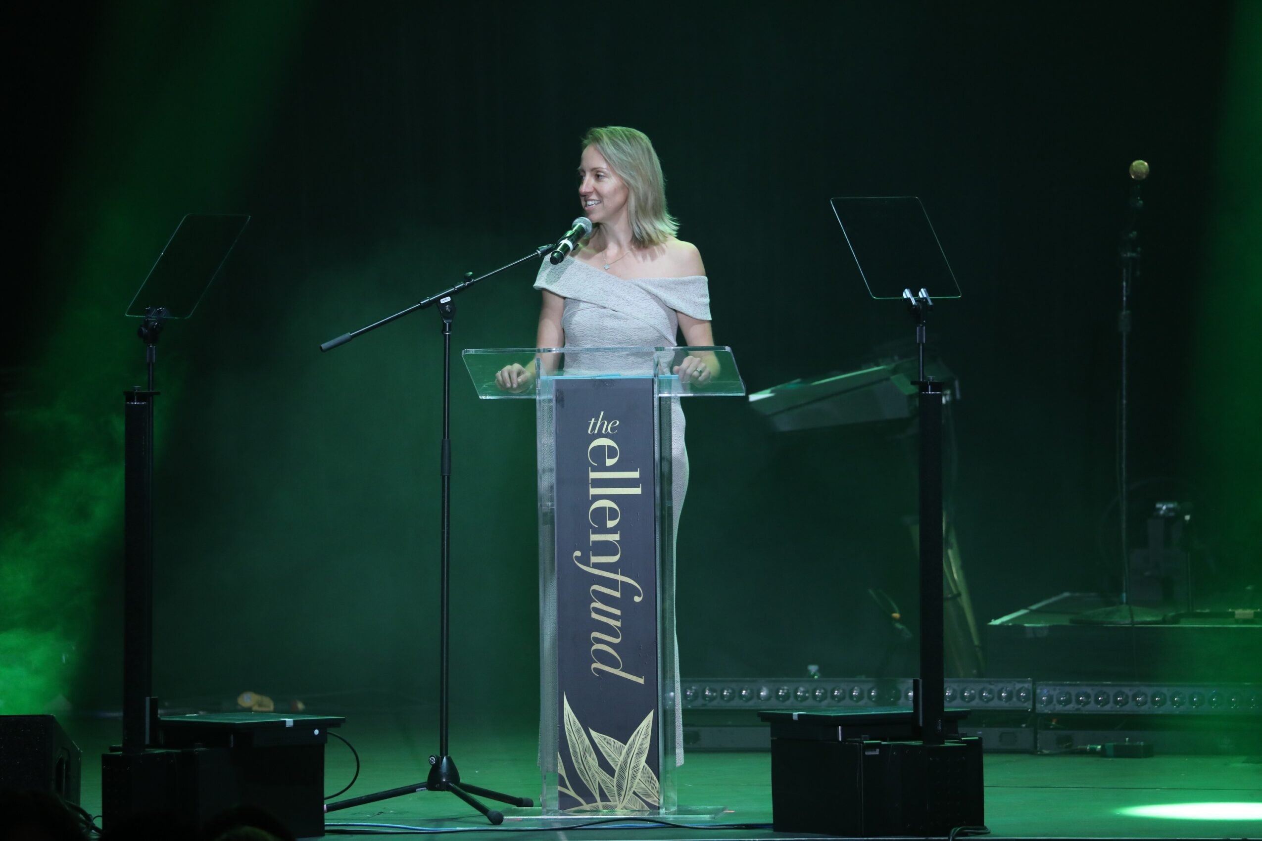 Woman standing at a transparent podium on stage with green lighting, speaking into a microphone.