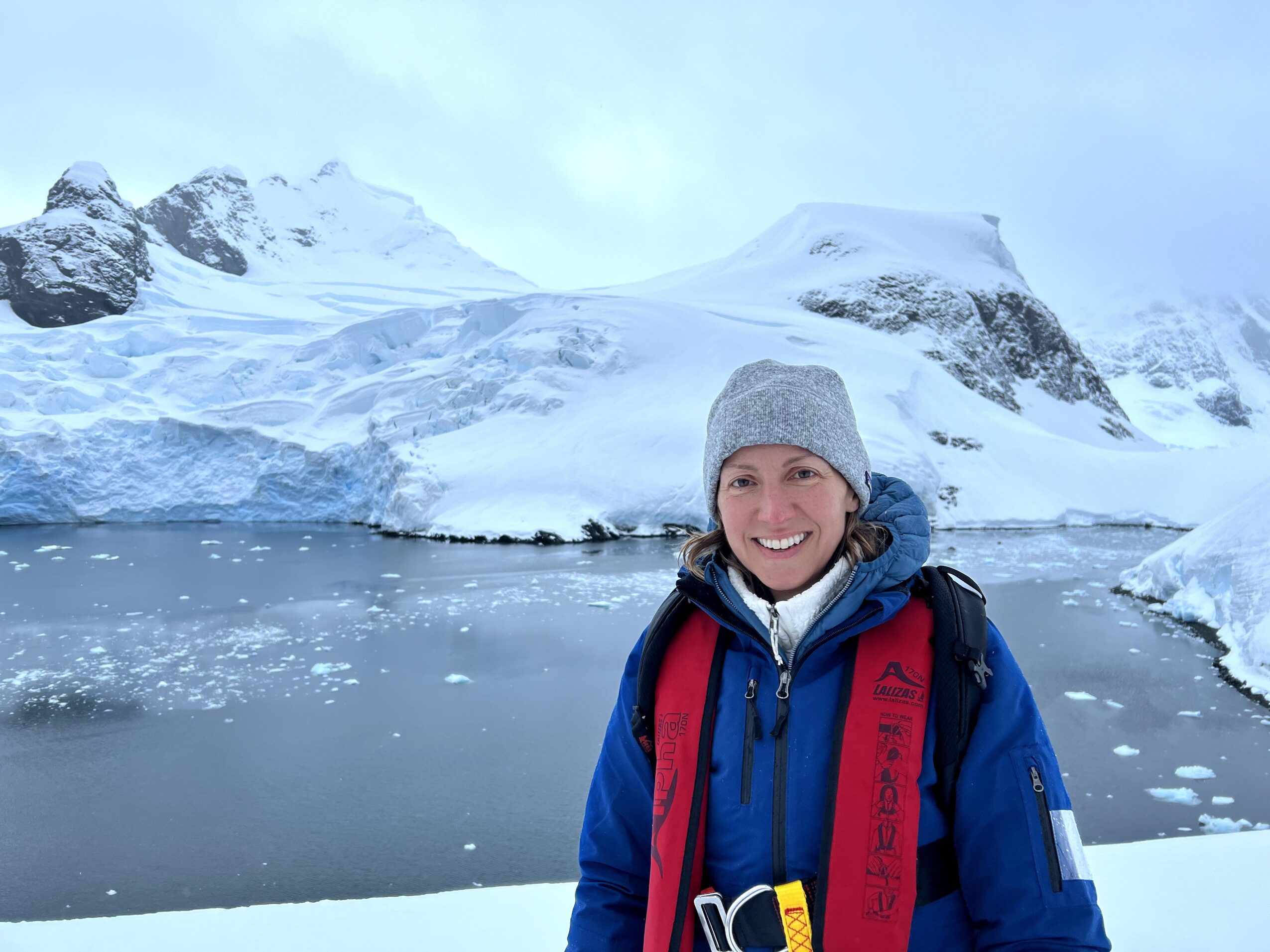 Woman in winter clothing and backpack smiling in icy landscape with snow-covered mountains and water