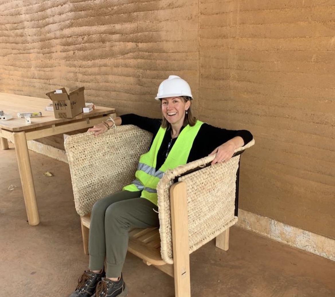 Woman in safety vest and helmet sitting on a chair in a construction site, smiling, with a wooden wall behind her.