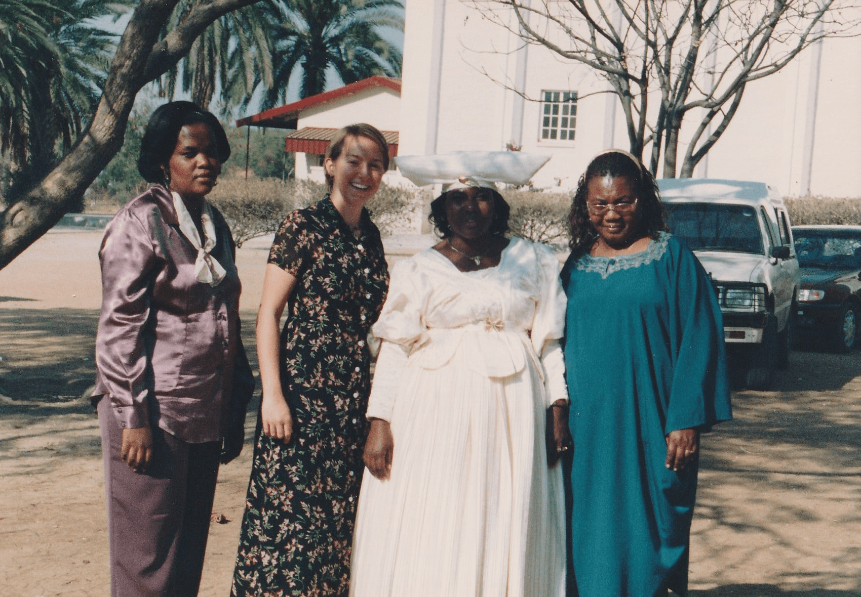 Four women standing outdoors, smiling, with trees and parked cars in the background.
