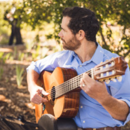 Man playing guitar outdoors with trees and sunlight in background.