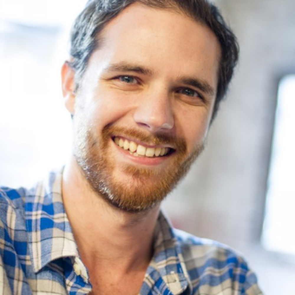 Smiling man with short dark hair and beard, wearing a blue plaid shirt, indoors with natural light.