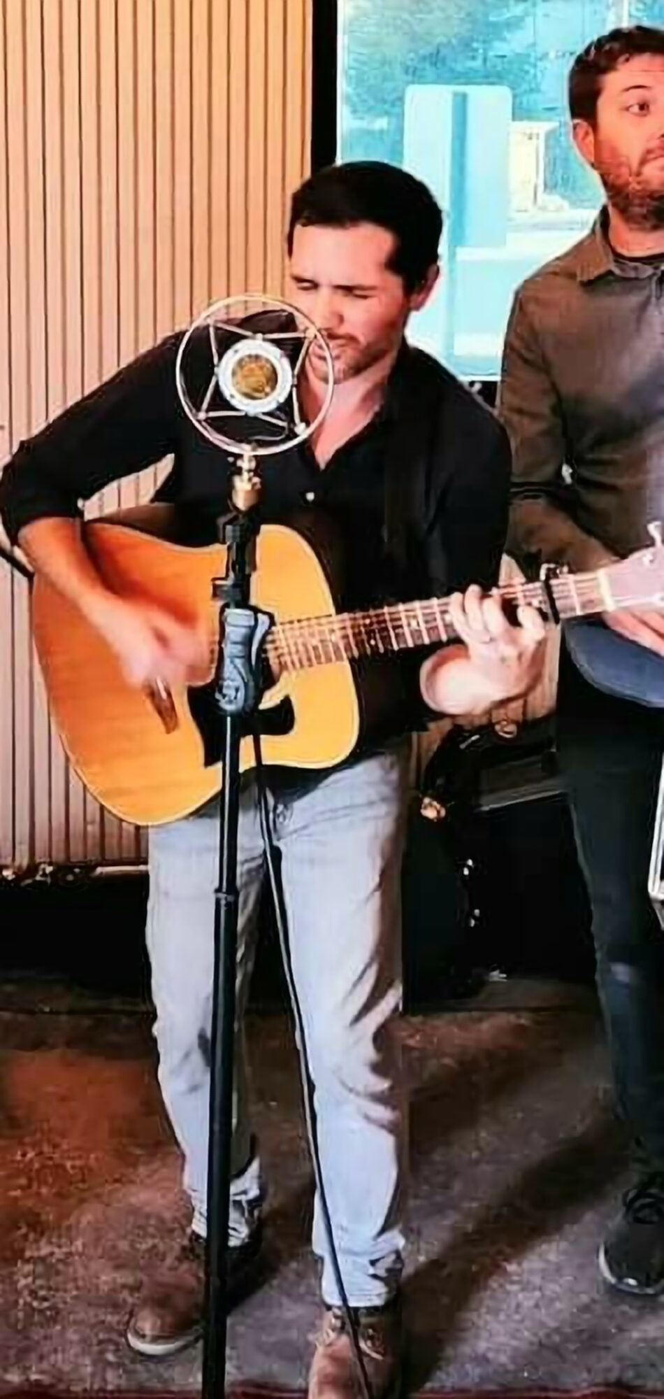 Person playing acoustic guitar with microphone, two others partially visible, indoor setting with window and wooden wall