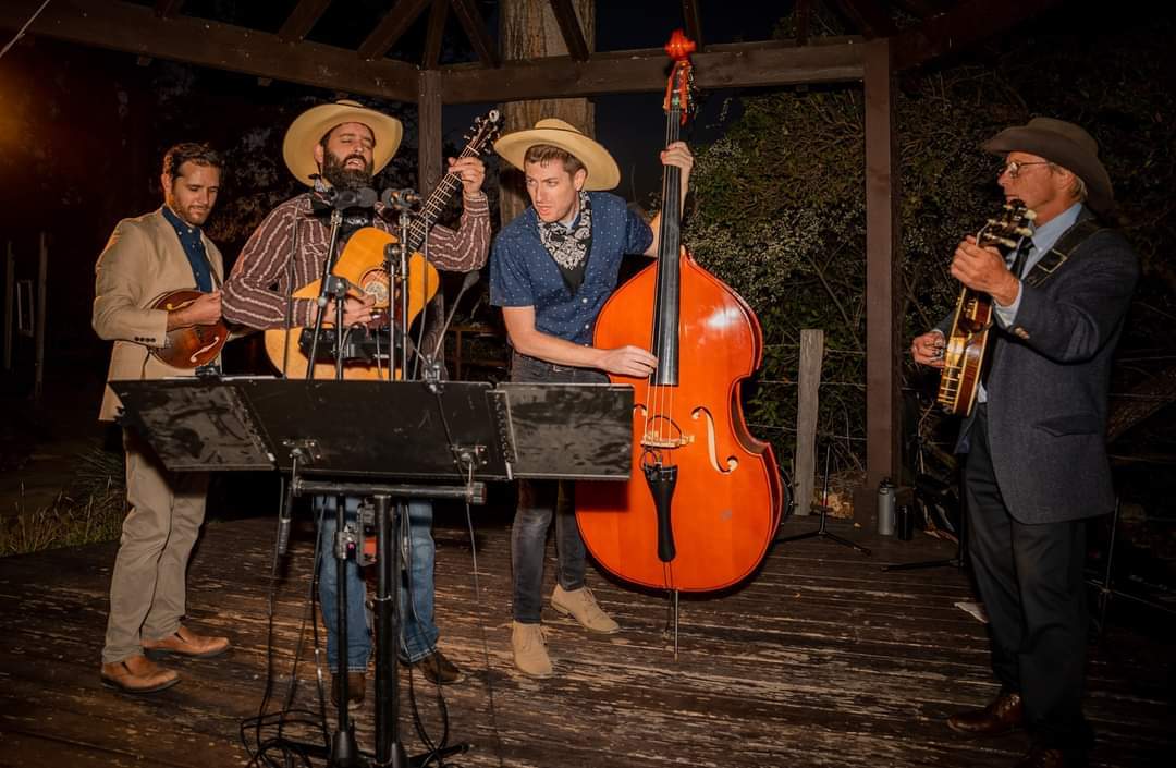 Four musicians perform on stage with guitars and a double bass, wearing hats and suits, under warm lighting.