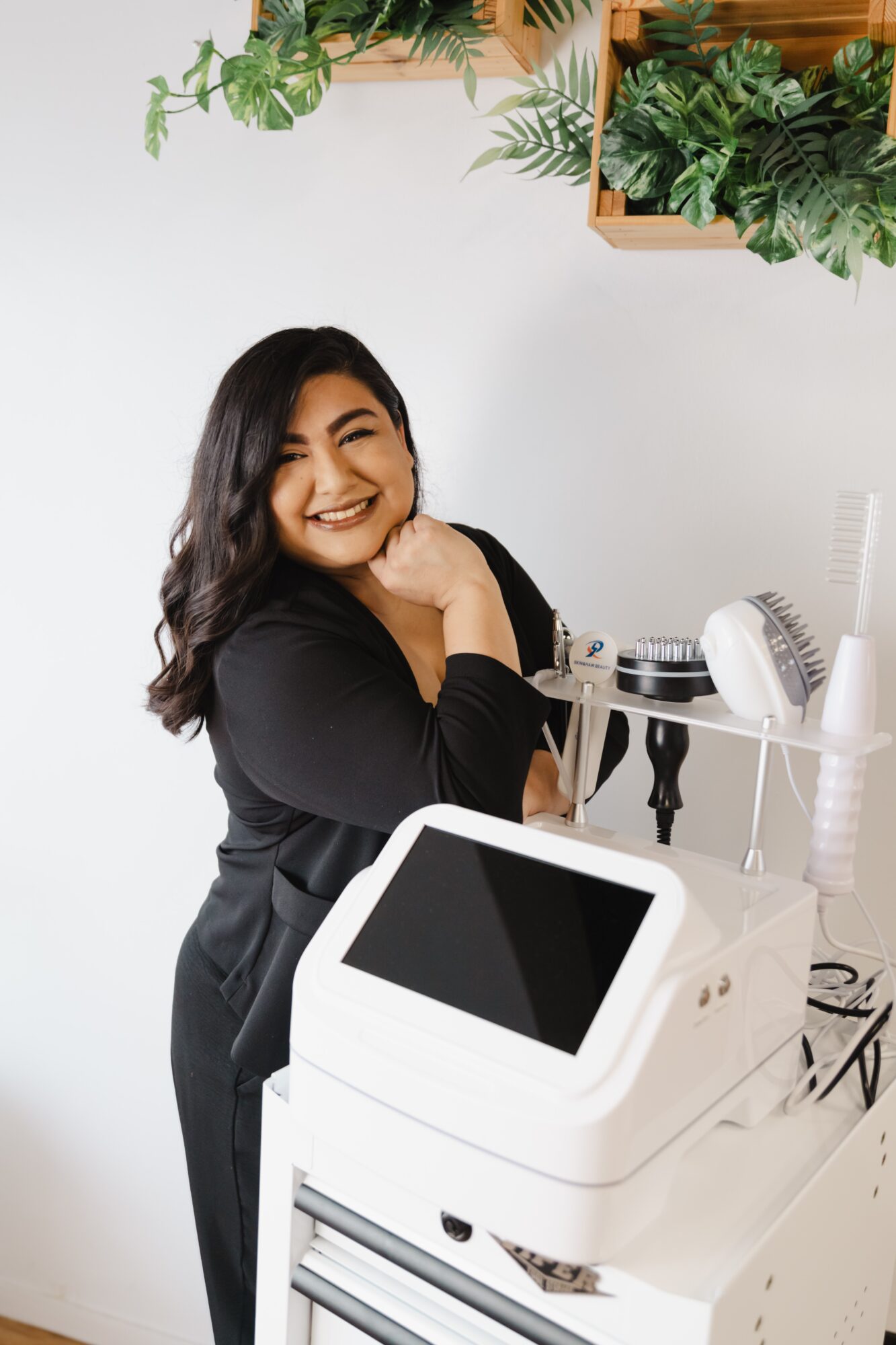 Smiling woman with dark hair in black outfit standing next to a medical or beauty device, with green plants above.