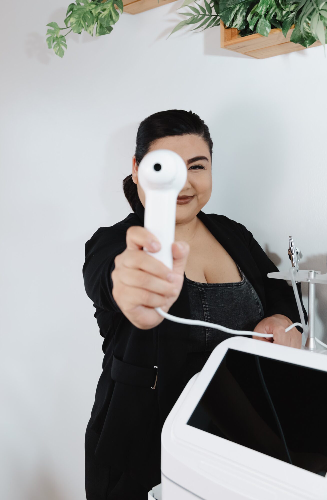 Woman holding a medical device towards the camera, smiling, with plants on the wall behind her.