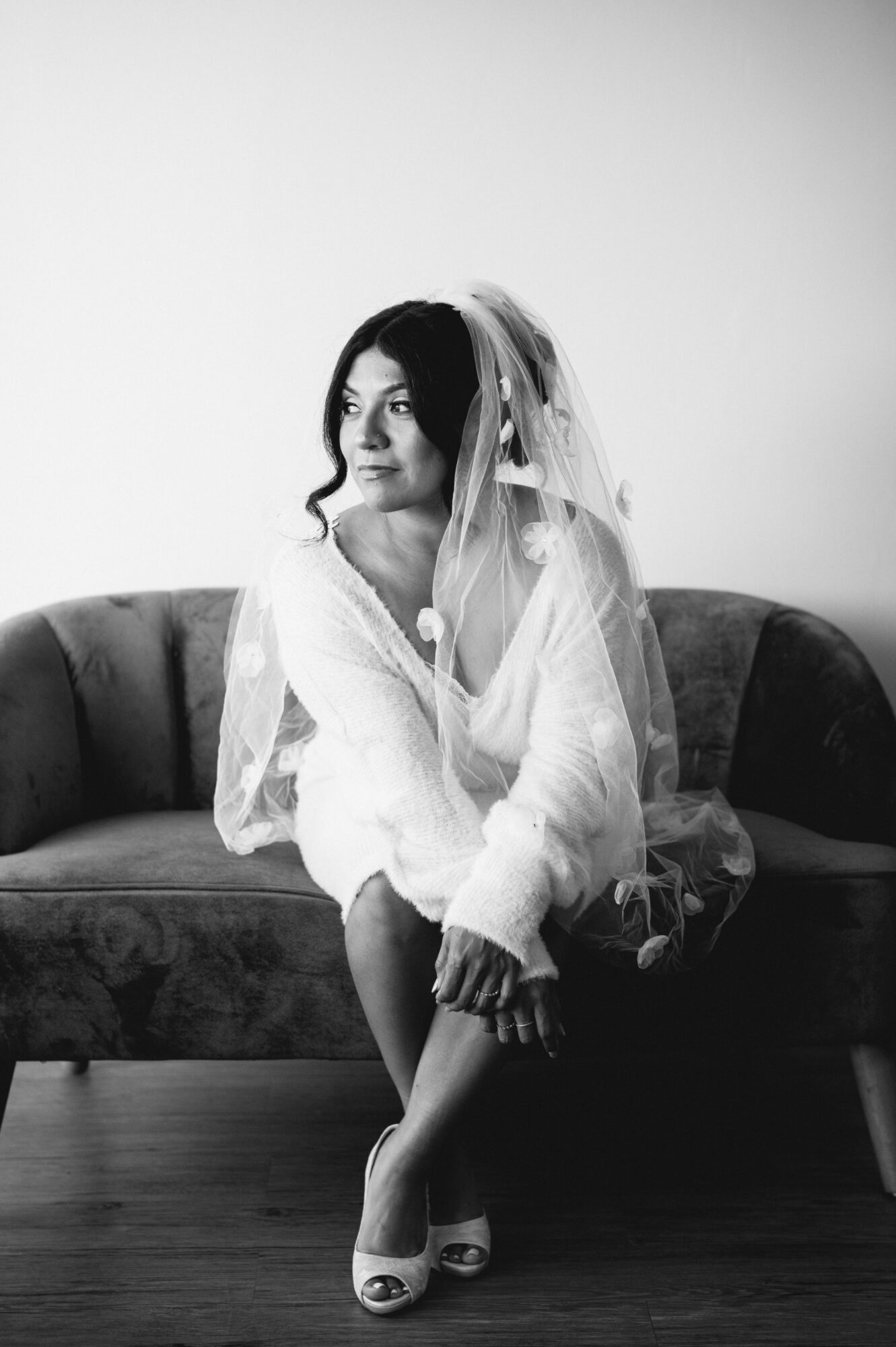 Woman sitting on a sofa wearing a veil and dress, looking to the side, in black and white.