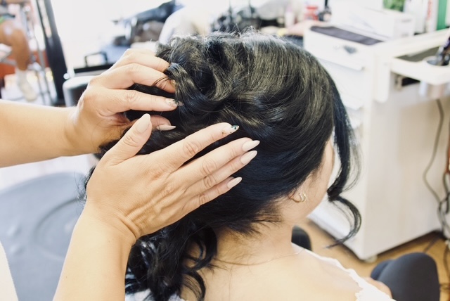 Person's head being styled with hands in dark, wavy hair, in a salon setting, with hair tools and equipment visible.