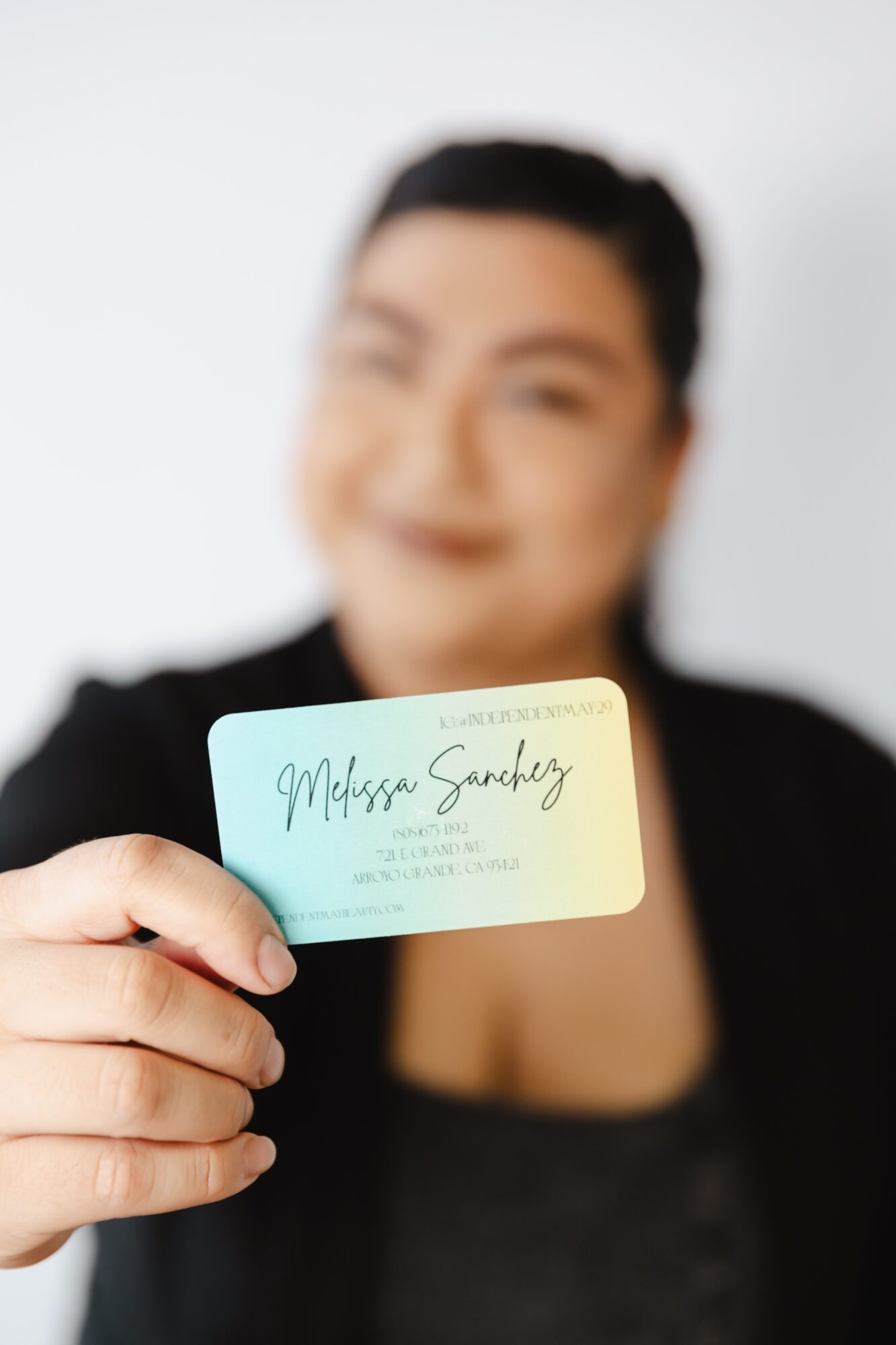 Person holding a business card with name and contact details, face blurred, background plain white.
