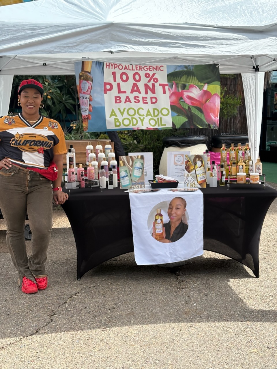 Child standing next to a table with bottles and a banner advertising plant-based avocado body oil, under a white canopy.