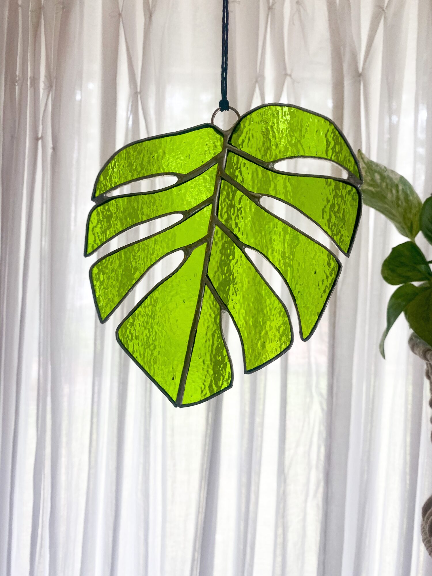 Green leaf-shaped stained glass hanging decoration with cut-out sections, hanging in front of a sheer curtain.
