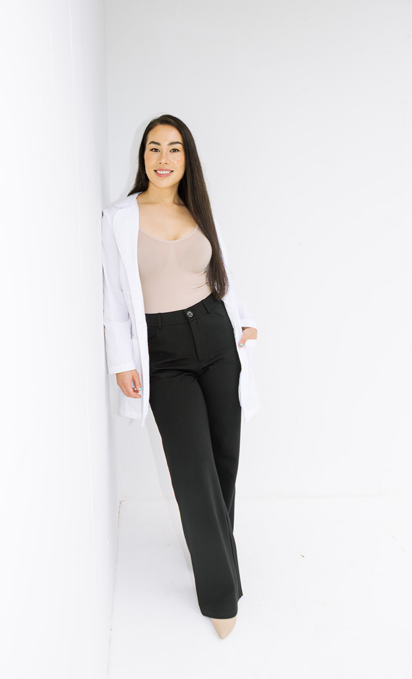 Woman with long dark hair leaning against a white wall, wearing a light top, black pants, and a white cardigan.