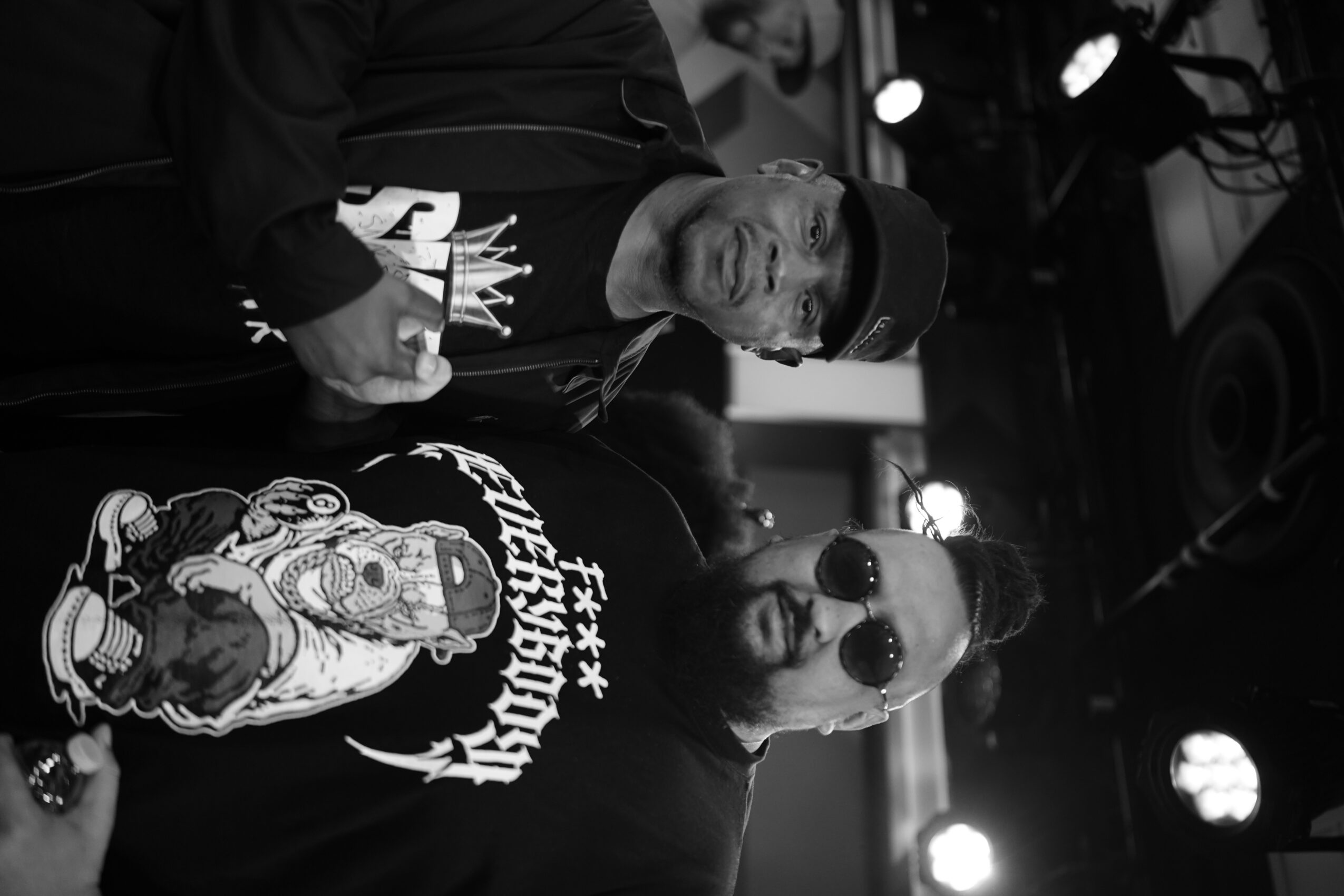 Two men, one wearing sunglasses and a hoodie, the other in a cap, pose indoors with a dark background.