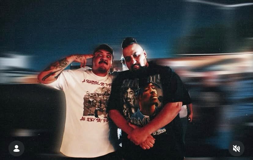 Two men stand close together, one with a tattooed arm, in a blurred background, facing the camera.