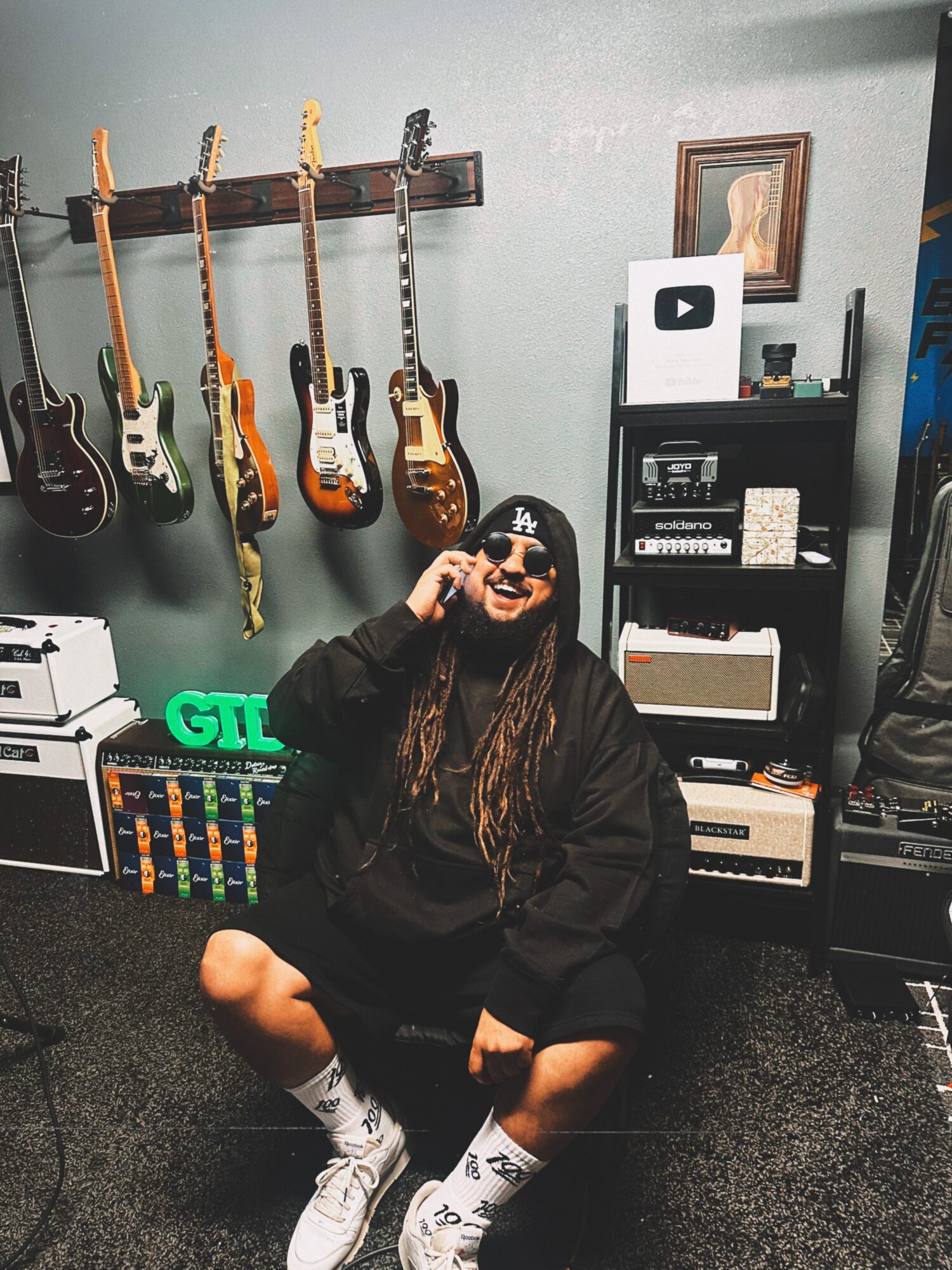 Person crouching in music studio with guitars on wall and equipment behind, smiling and wearing sunglasses.