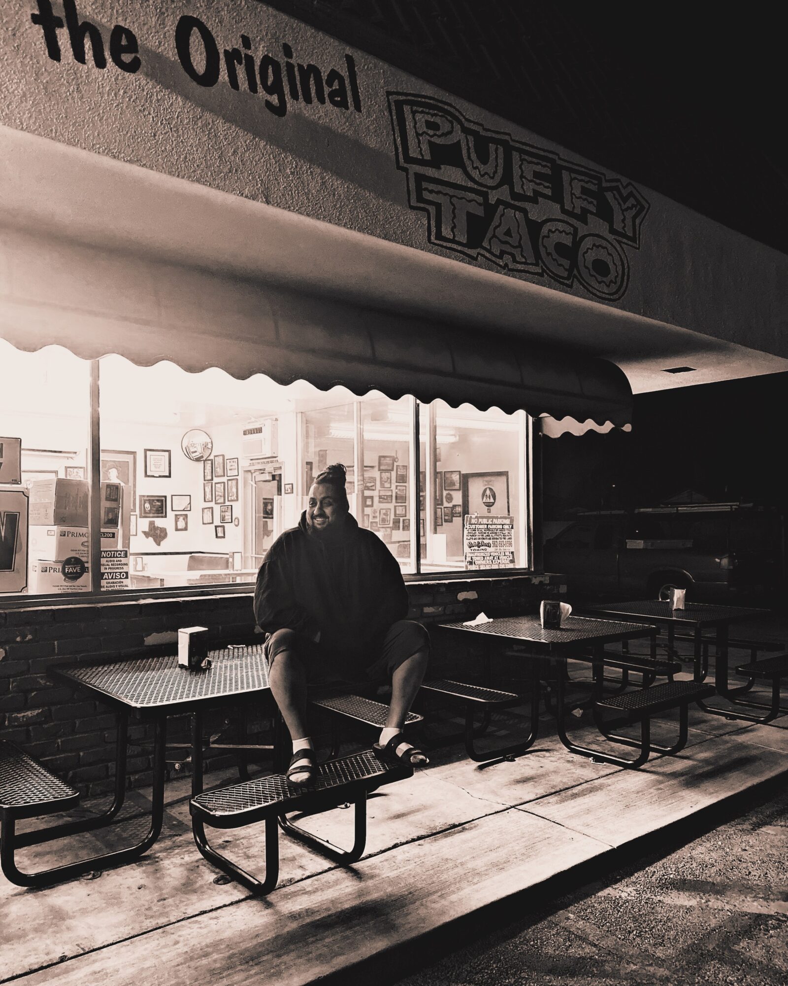 Person sitting on a bench outside a taco restaurant at night, with illuminated interior visible through large windows.