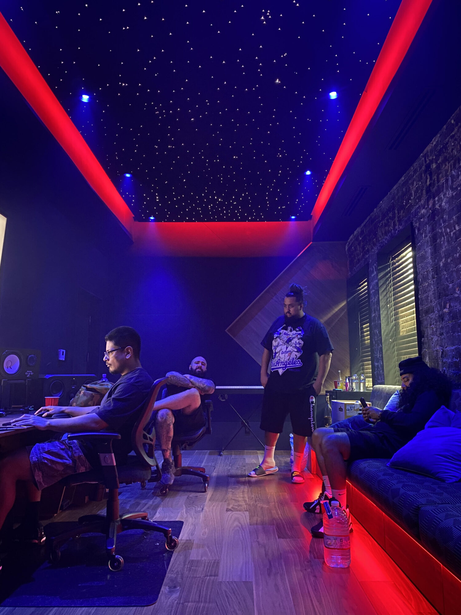 People in a dimly lit room with a starry ceiling, red lighting, and large windows, some working at desks and others seated.