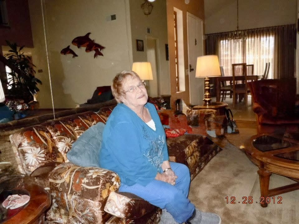 Older woman with glasses and short hair sitting on a patterned sofa in a living room, smiling, with lamps and curtains in background.