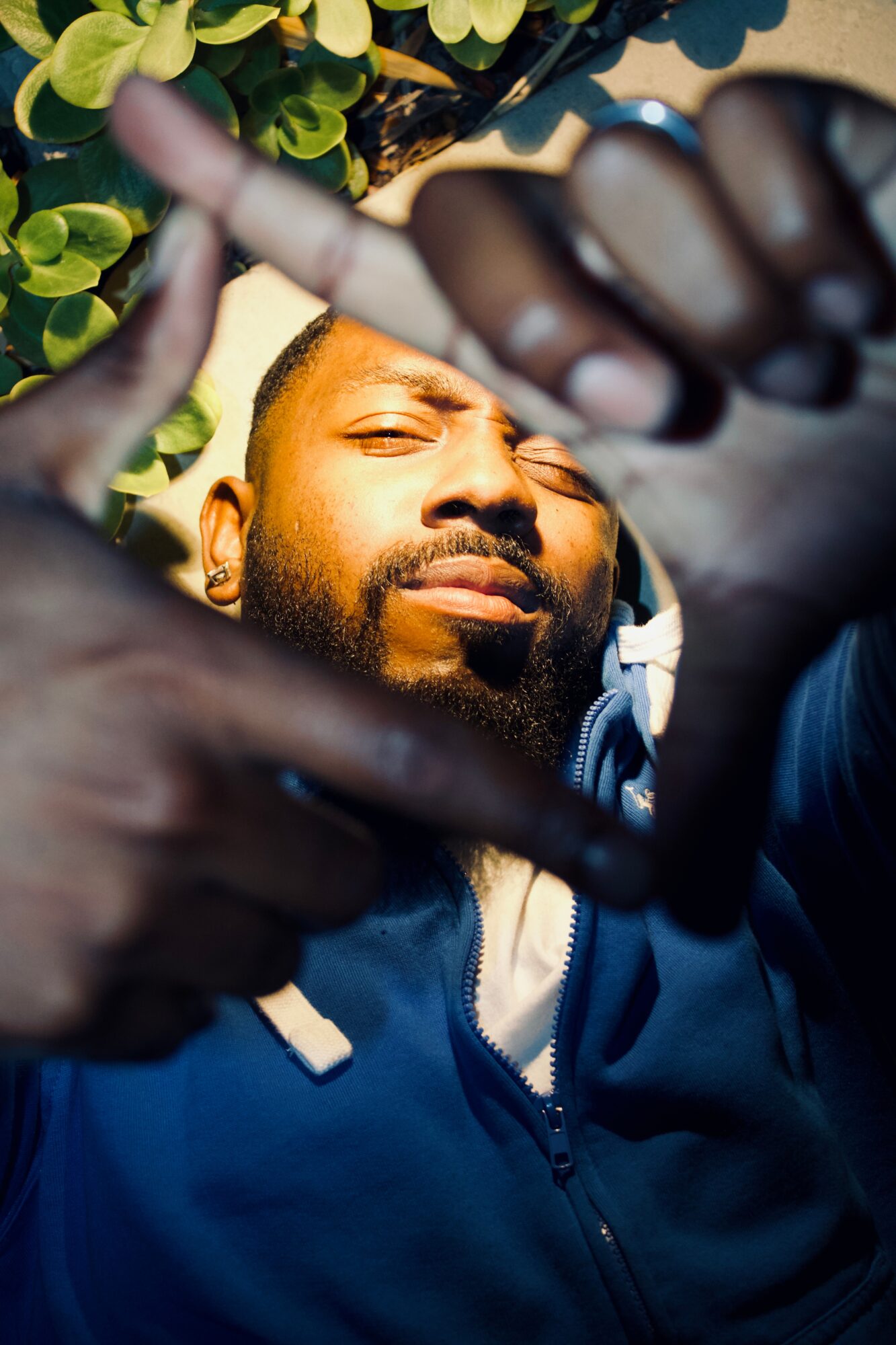 Man lying outdoors, eyes closed, surrounded by greenery, with hands forming a frame around his face.
