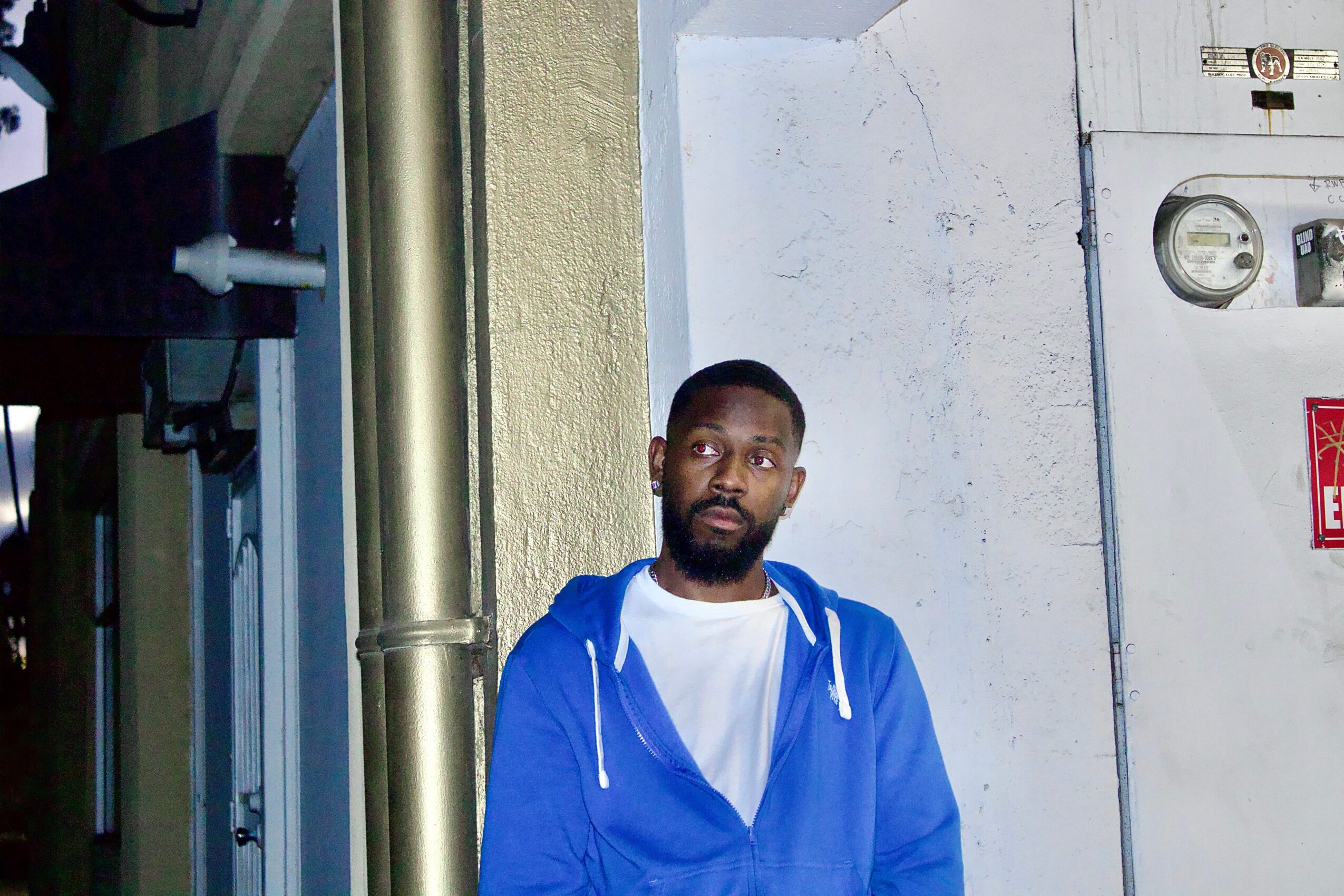 Man with a beard wearing a blue hoodie and white t-shirt standing outdoors against a wall.