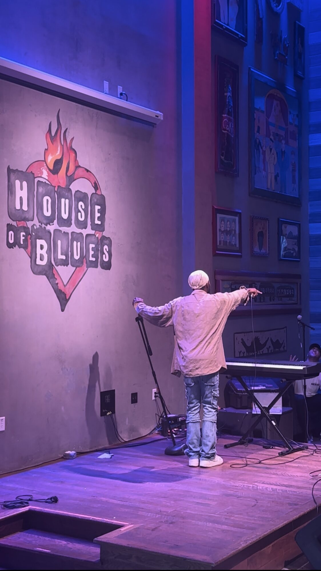 Person on stage with arms outstretched, facing a wall with a 'House of Blues' logo, in a dimly lit venue.