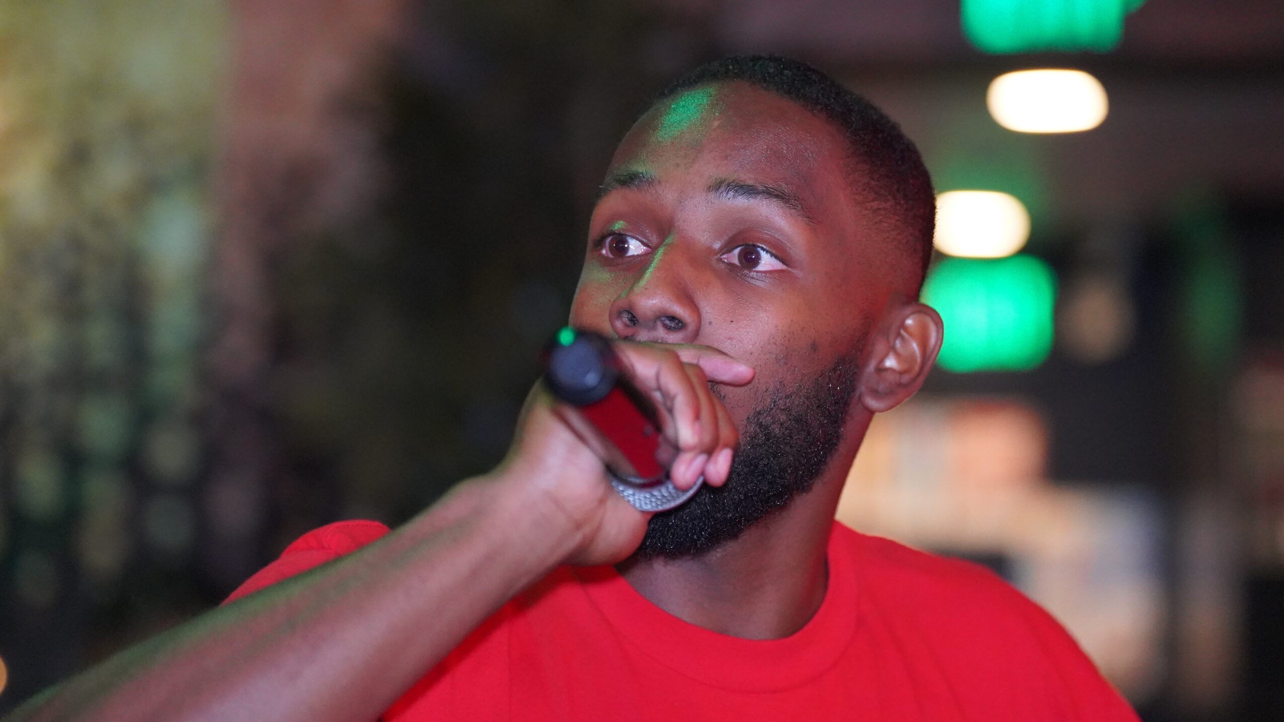Man singing into a microphone with a surprised expression, wearing a red shirt, in a dimly lit environment.