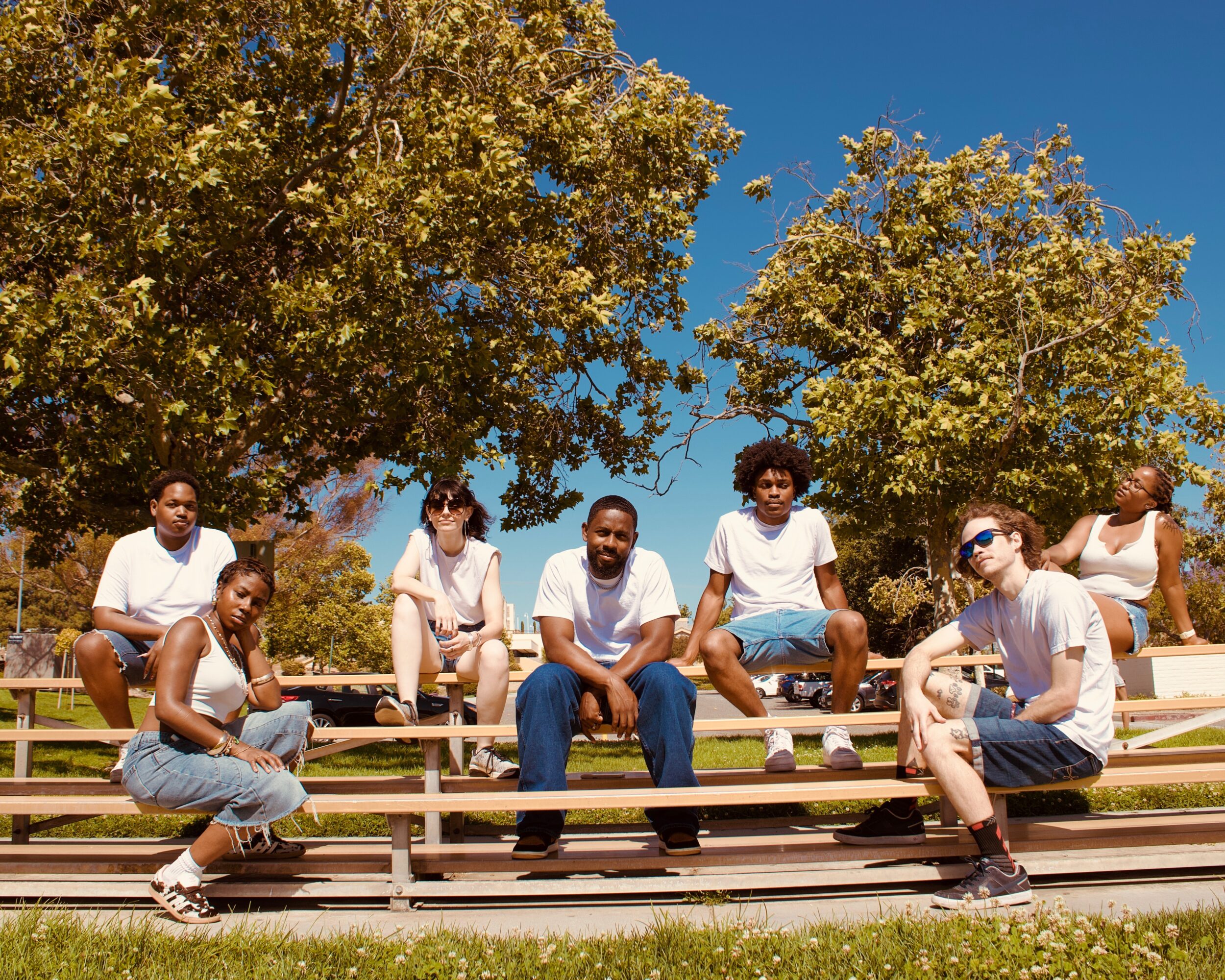 Group of six people sitting on bleachers outdoors under trees with green leaves and blue sky.