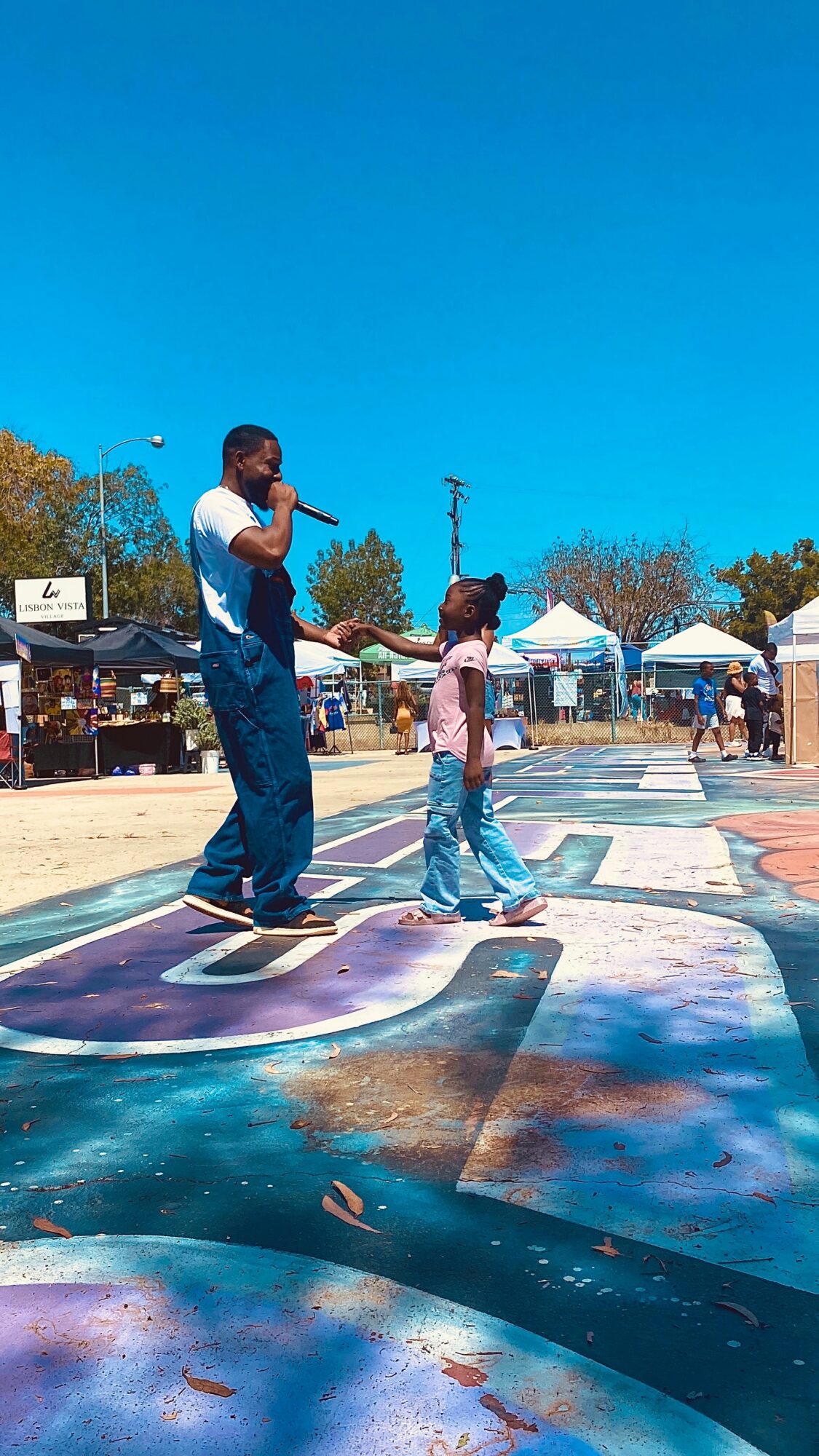 A man and a girl stand on colorful painted pavement outdoors under a clear blue sky, with tents and people in the background.