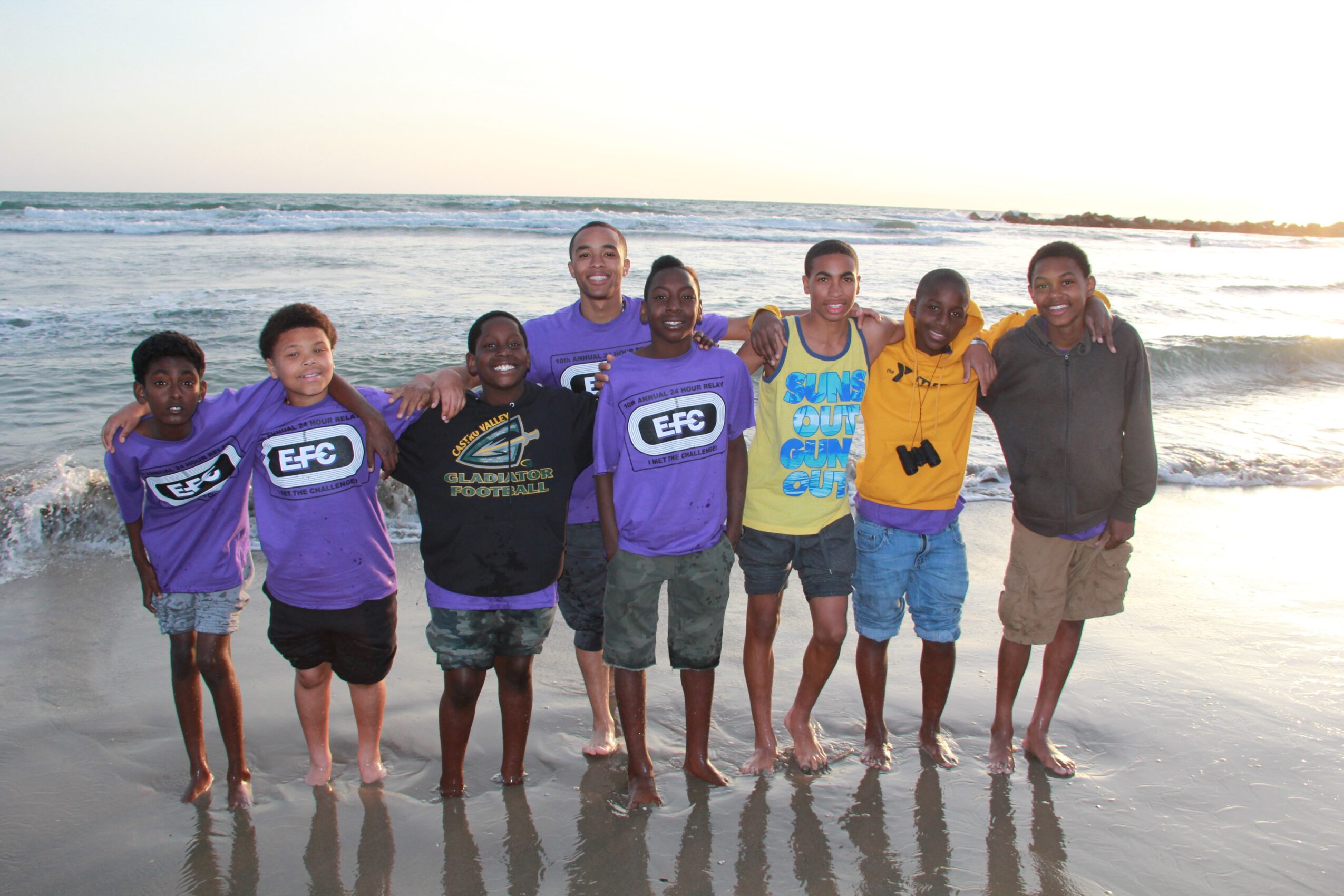 Group of children and a young adult standing on the beach with ocean waves behind them, smiling, some with arms around each other.