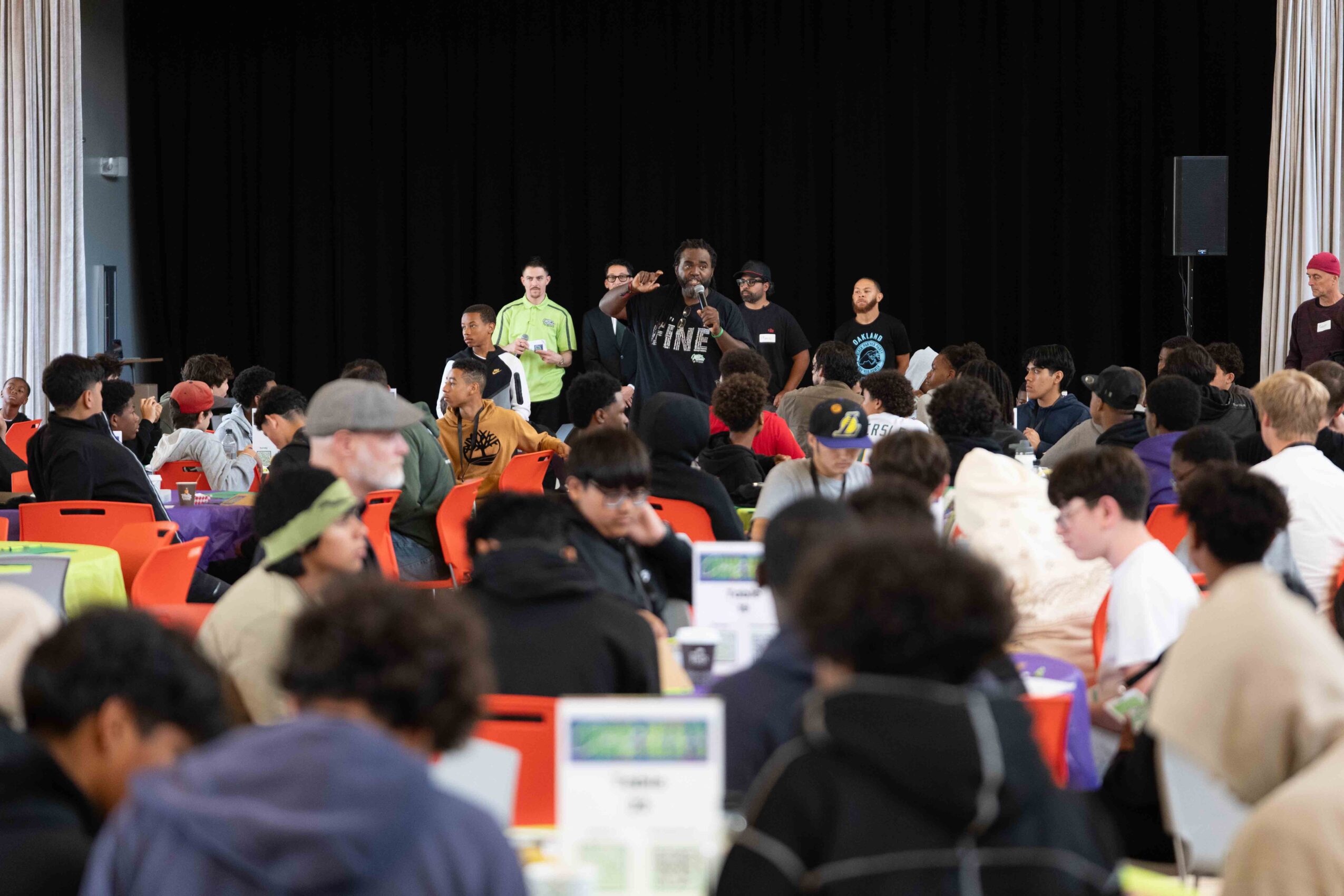 A speaker addresses a large audience in a hall with a black curtain backdrop, people seated at tables, some using laptops.
