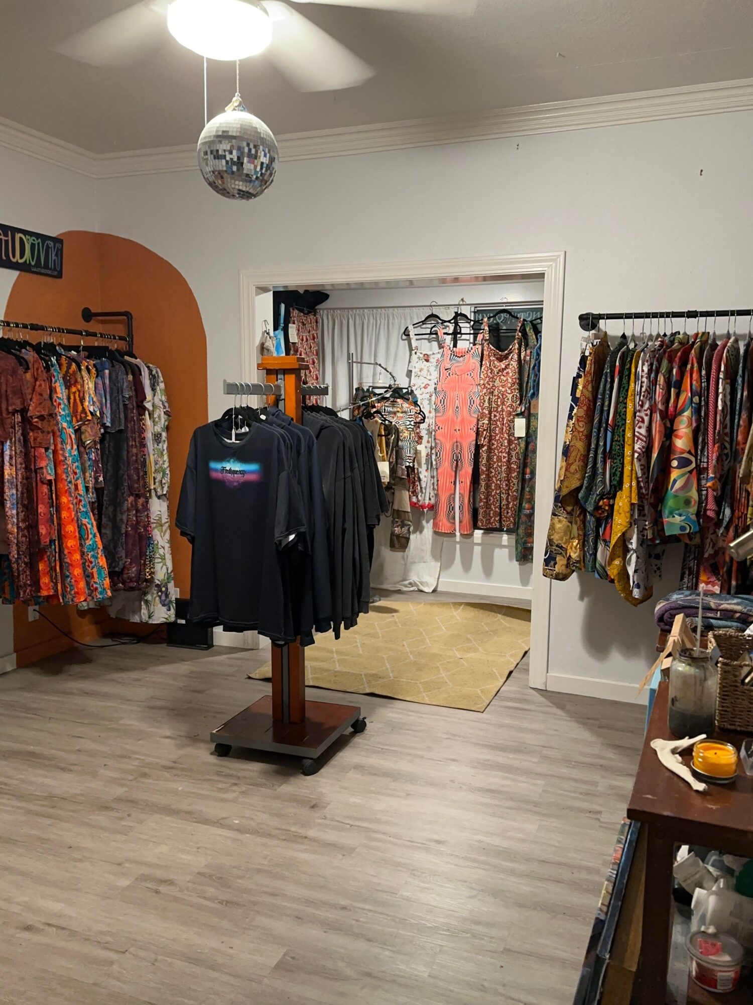 Clothing store with racks of colorful garments, a black jacket on a stand, and a wooden table, with a mirror and ceiling light.