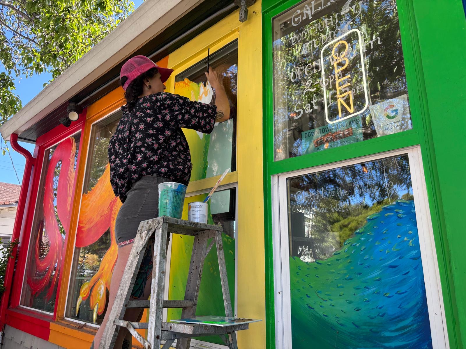 Person painting a colorful mural on a building exterior, wearing a pink hat and standing on a ladder.