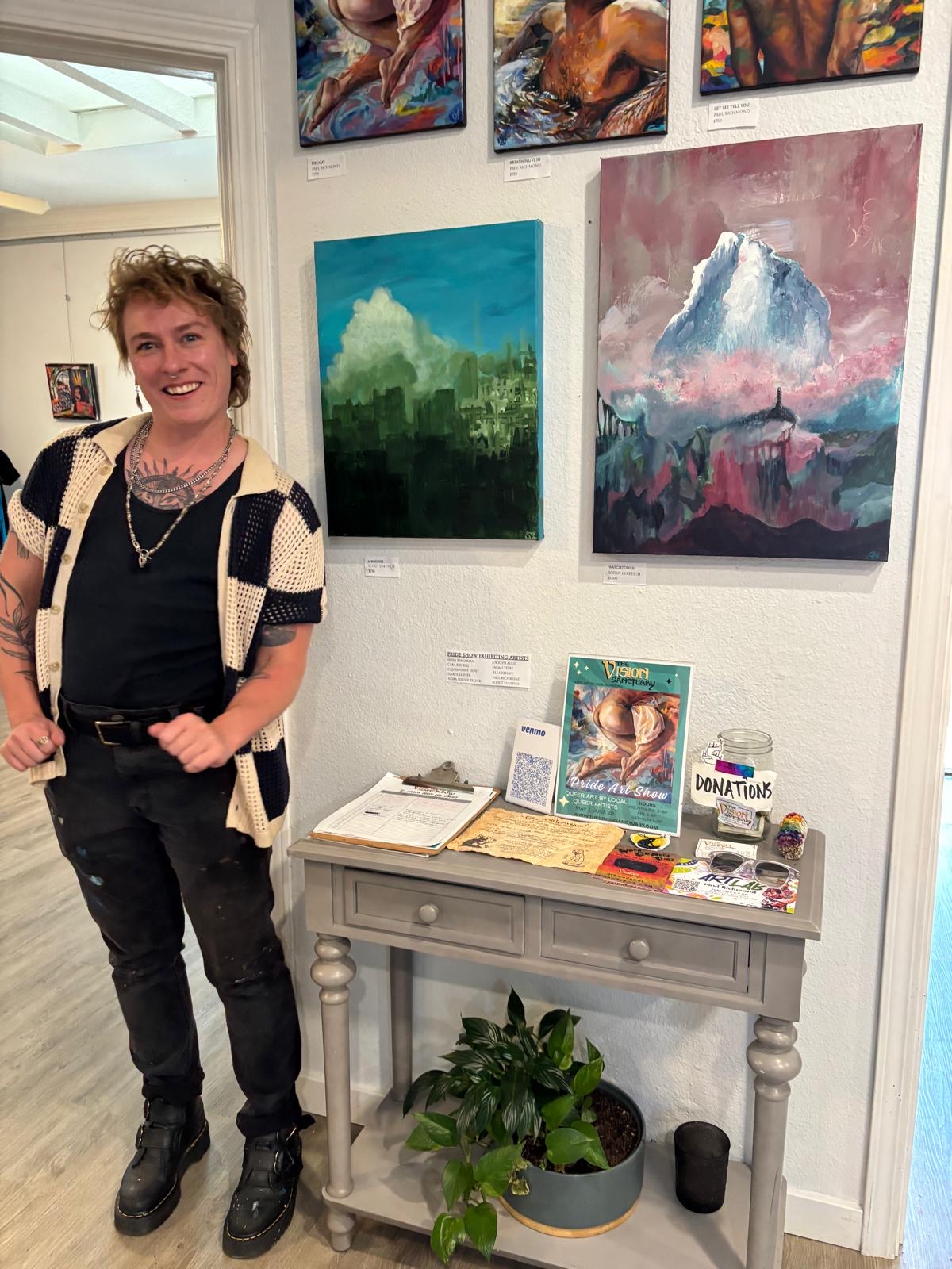 Person with short curly hair smiling, wearing a black shirt, patterned cardigan, and black pants, standing in an art gallery.