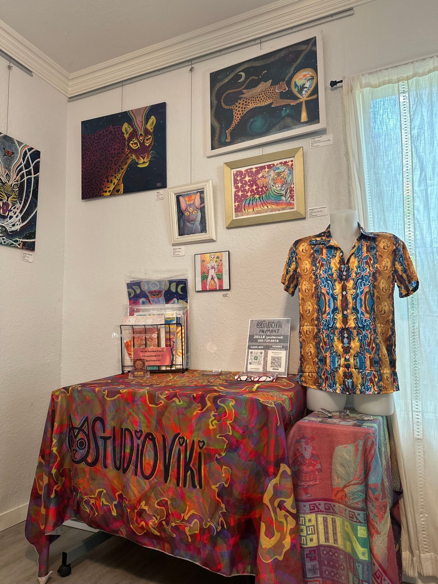 Room corner with colorful fabric-covered table, wall art, and a window with curtains, viewed from the side.