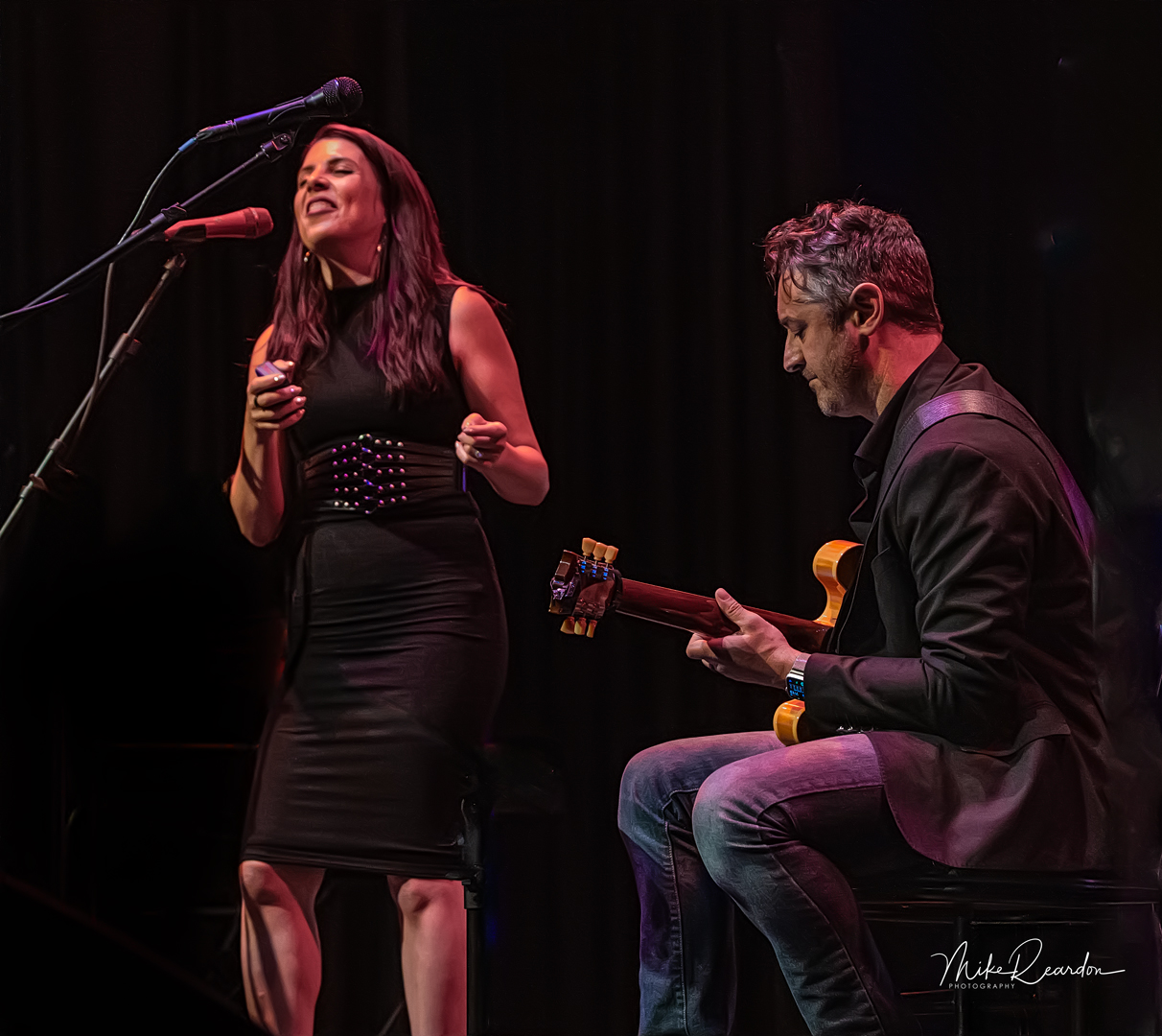 A woman singing with a microphone and a man playing guitar on stage, both engaged in performance, dark background.