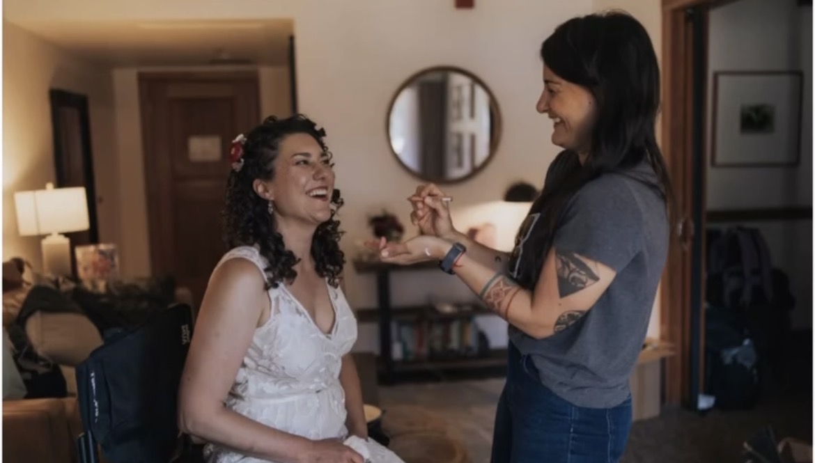 Two women smiling and interacting indoors, one sitting and the other standing, in a cozy room with warm lighting.