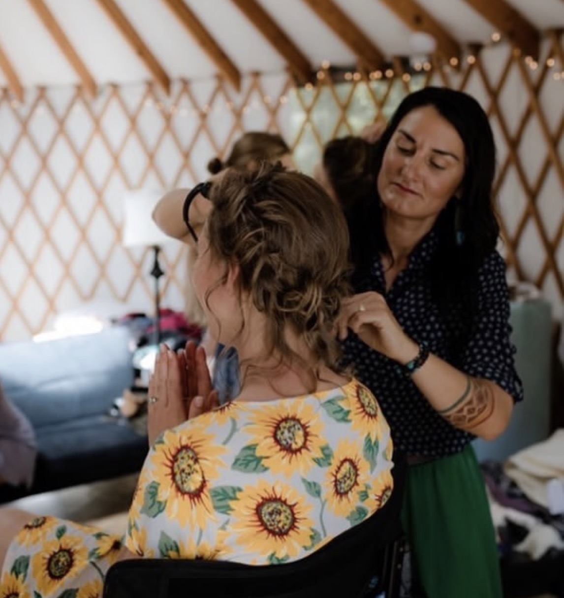 Woman with dark hair styles a woman's hair inside a yurt, with a sunflower-patterned shirt, in a cozy setting.