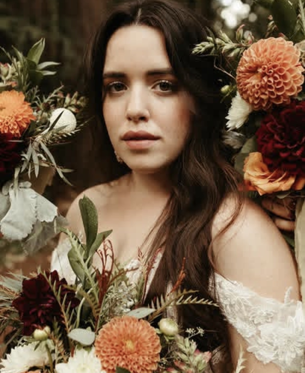 Young woman with long dark hair surrounded by colorful flowers, looking directly at the camera.