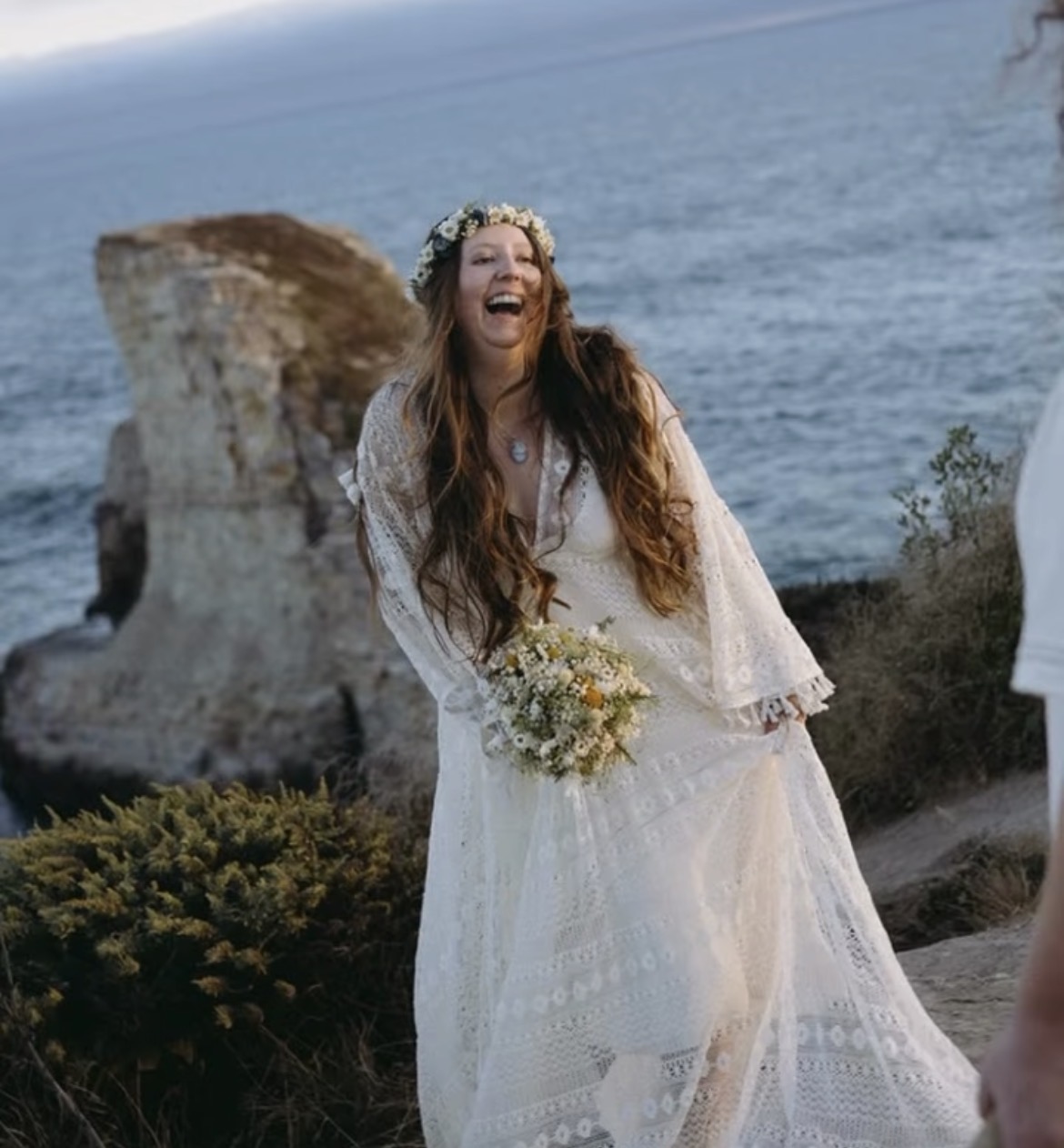 Woman in a white dress with a flower crown, holding a bouquet, laughing outdoors near the sea with a rock formation in the background.