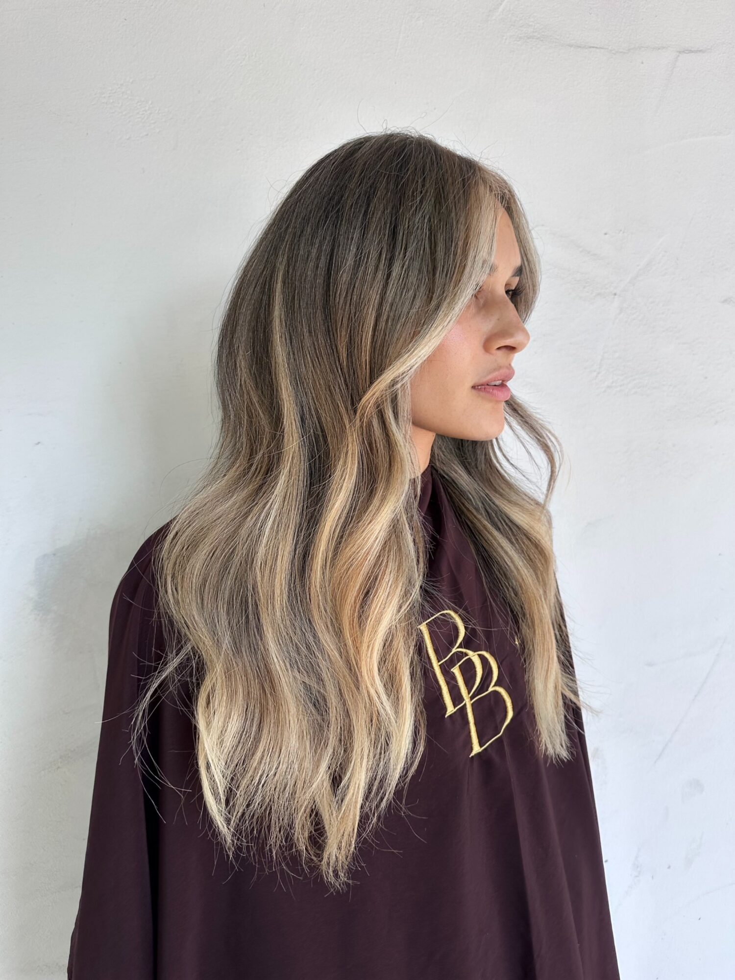 Side profile of a woman with long, wavy blonde hair wearing a dark top with gold lettering, standing against a white wall.
