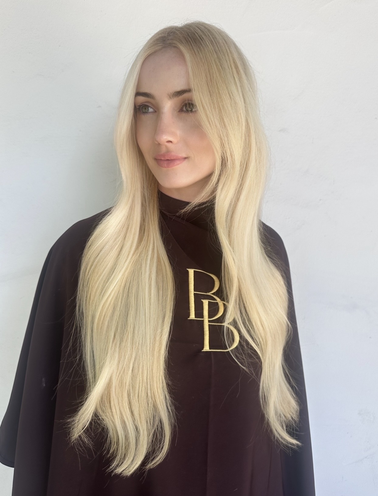Young woman with long blonde hair wearing a black top with gold lettering, standing against a white background.