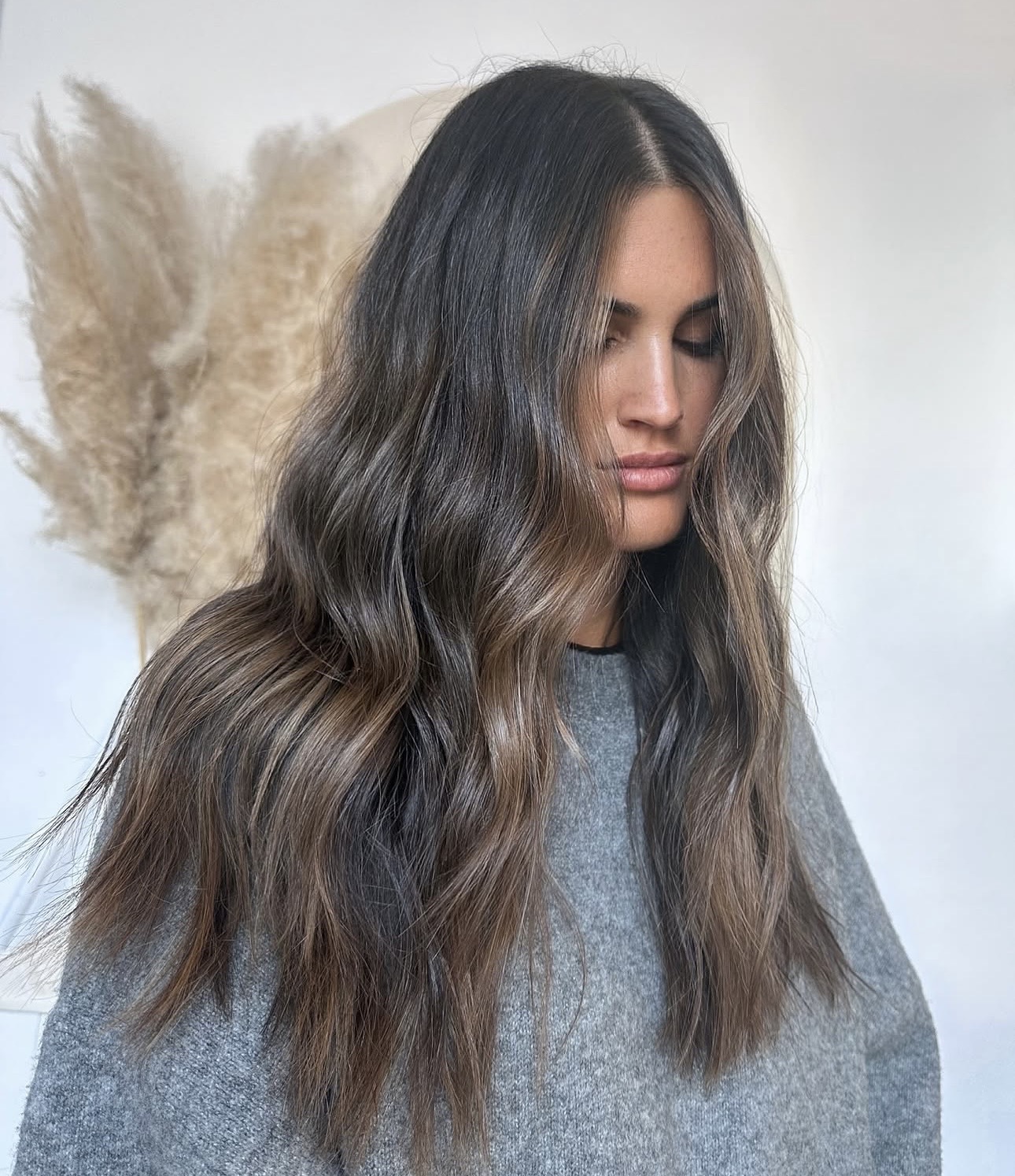 Woman with long wavy hair and gray shirt, neutral background, looking down slightly.